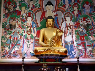 A golden statue of Buddha sits serenely in the foreground, surrounded by an ornate and colorful mural depicting various deities and figures. The statue is adorned in traditional robes and positioned in the center, while the mural includes intricate details with vibrant colors and elaborate patterns. The scene conveys a sense of spirituality and reverence.