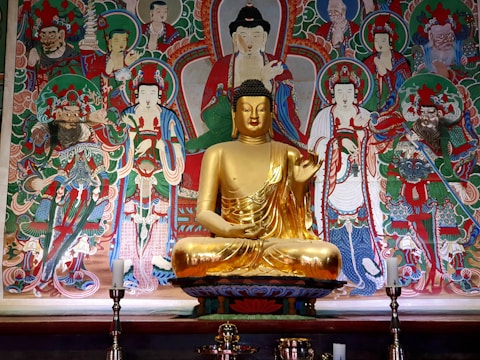 A golden statue of Buddha sits serenely in the foreground, surrounded by an ornate and colorful mural depicting various deities and figures. The statue is adorned in traditional robes and positioned in the center, while the mural includes intricate details with vibrant colors and elaborate patterns. The scene conveys a sense of spirituality and reverence.