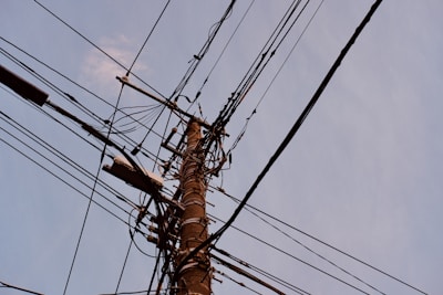 A wooden utility pole is surrounded by numerous intersecting power lines against a backdrop of a clear sky. The pole is equipped with various insulators and connectors, creating a complex network of cables.