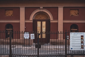 An entrance to a historic-looking building with a locked metal gate in the foreground. The building features two bas-relief sculptures and is constructed with a reddish-brown facade and beige accents. Signs on the gate provide information and entry instructions in multiple languages.