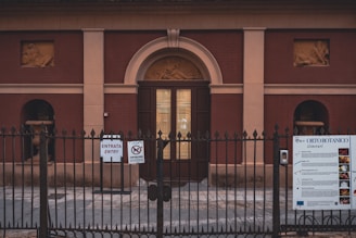 An entrance to a historic-looking building with a locked metal gate in the foreground. The building features two bas-relief sculptures and is constructed with a reddish-brown facade and beige accents. Signs on the gate provide information and entry instructions in multiple languages.