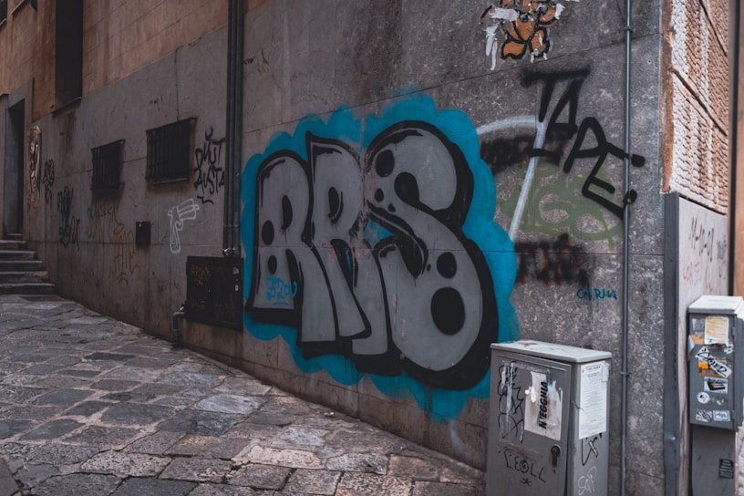 An urban alleyway features a prominent graffiti piece on a stone wall with the letters 'BRS' in bold black and white, surrounded by a blue outline. The surrounding walls display various smaller graffiti tags and stickers. The ground is paved with uneven stone tiles, and a few utility boxes are present, also covered in stickers and graffiti.