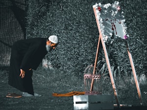 A man is bending over in a garden, wearing a black robe and a white cap. In front of him, there is a decorative standing sign adorned with flowers. Beside him on the grass are a pair of brown shoes and a folded piece of cloth.