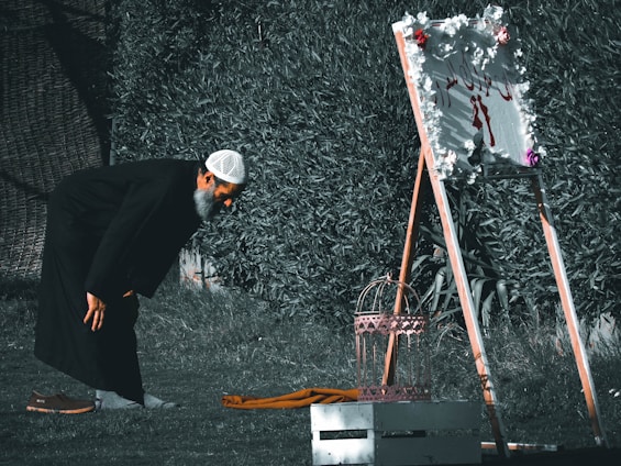 A man is bending over in a garden, wearing a black robe and a white cap. In front of him, there is a decorative standing sign adorned with flowers. Beside him on the grass are a pair of brown shoes and a folded piece of cloth.