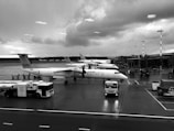 Several planes are parked at an airport terminal on a rainy day. The aircraft have 'airBaltic' branding visible on the fuselage. There are service vehicles and equipment around the airplanes on the wet tarmac. Raindrops are visible on the window through which the photo is taken.