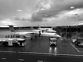 Several planes are parked at an airport terminal on a rainy day. The aircraft have 'airBaltic' branding visible on the fuselage. There are service vehicles and equipment around the airplanes on the wet tarmac. Raindrops are visible on the window through which the photo is taken.