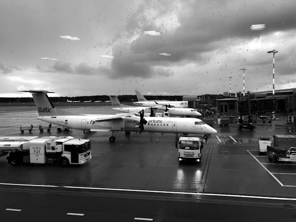 Several planes are parked at an airport terminal on a rainy day. The aircraft have 'airBaltic' branding visible on the fuselage. There are service vehicles and equipment around the airplanes on the wet tarmac. Raindrops are visible on the window through which the photo is taken.