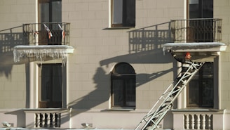 A firefighter wearing a helmet and protective gear is positioned on a ladder leaning against a building facade. The structure features two balconies with shadows cast by the railings. The firefighter appears to be reaching towards the icicles hanging from one of the balconies.