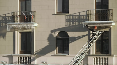A firefighter wearing a helmet and protective gear is positioned on a ladder leaning against a building facade. The structure features two balconies with shadows cast by the railings. The firefighter appears to be reaching towards the icicles hanging from one of the balconies.