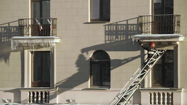 A firefighter wearing a helmet and protective gear is positioned on a ladder leaning against a building facade. The structure features two balconies with shadows cast by the railings. The firefighter appears to be reaching towards the icicles hanging from one of the balconies.