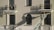 A firefighter wearing a helmet and protective gear is positioned on a ladder leaning against a building facade. The structure features two balconies with shadows cast by the railings. The firefighter appears to be reaching towards the icicles hanging from one of the balconies.