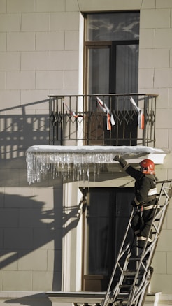 A firefighter stands on a ladder reaching up to clear long icicles hanging from a building's balcony railing. The building facade is light-colored with visible shadows, and the balcony is decorated with red and white ribbons.