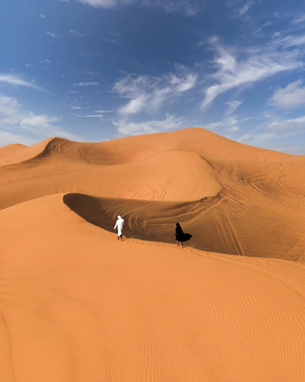 A man in a black jacket and white trousers standing on sand under a blue sky