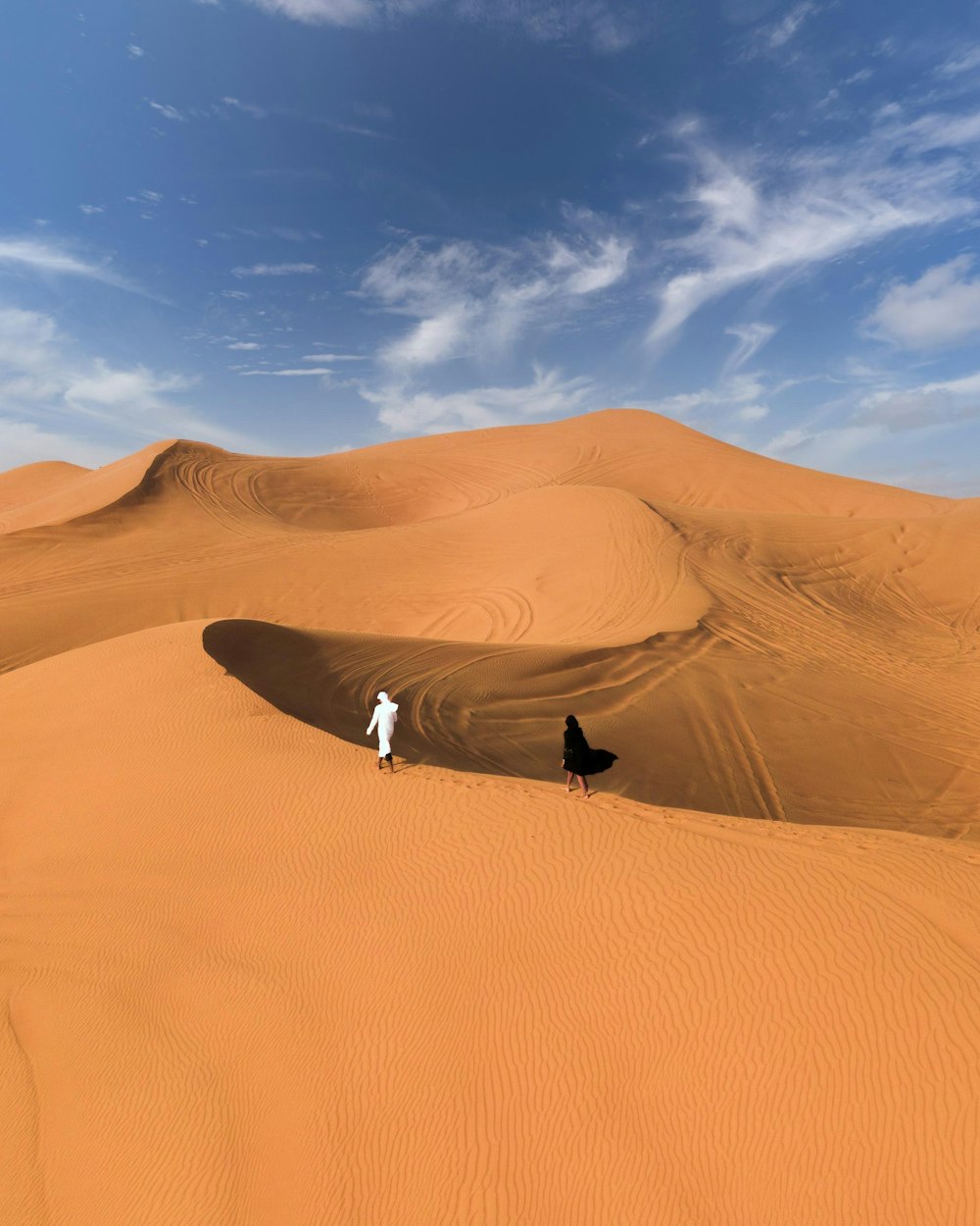 Mann in schwarzer Jacke und weißer Hose steht tagsüber auf braunem Sand unter blauem Himmel
