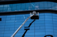 A facade with large blue-tinted glass windows dominates the scene. In front of the tall building, a hydraulic lift extends upwards with a work platform near the middle, likely used for window cleaning or maintenance.