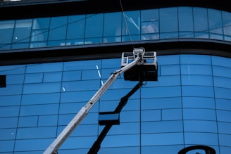 A facade with large blue-tinted glass windows dominates the scene. In front of the tall building, a hydraulic lift extends upwards with a work platform near the middle, likely used for window cleaning or maintenance.