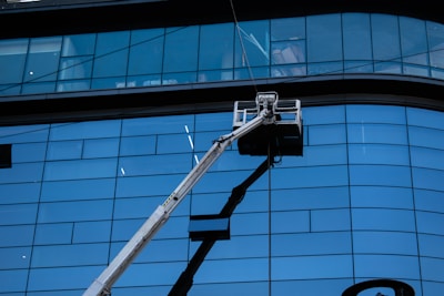 A facade with large blue-tinted glass windows dominates the scene. In front of the tall building, a hydraulic lift extends upwards with a work platform near the middle, likely used for window cleaning or maintenance.