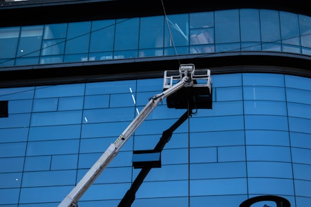 A facade with large blue-tinted glass windows dominates the scene. In front of the tall building, a hydraulic lift extends upwards with a work platform near the middle, likely used for window cleaning or maintenance.