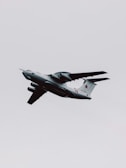 A military aircraft with distinctive radars and aerodynamic design flies through a cloudy sky. The airplane is painted in gray and has recognizable markings on its body, with four powerful jet engines under its wings.