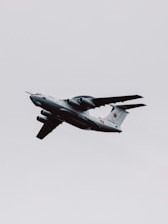 A military aircraft with distinctive radars and aerodynamic design flies through a cloudy sky. The airplane is painted in gray and has recognizable markings on its body, with four powerful jet engines under its wings.