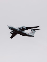 A military aircraft with distinctive radars and aerodynamic design flies through a cloudy sky. The airplane is painted in gray and has recognizable markings on its body, with four powerful jet engines under its wings.