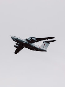 A military aircraft with distinctive radars and aerodynamic design flies through a cloudy sky. The airplane is painted in gray and has recognizable markings on its body, with four powerful jet engines under its wings.