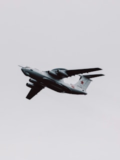 A military aircraft with distinctive radars and aerodynamic design flies through a cloudy sky. The airplane is painted in gray and has recognizable markings on its body, with four powerful jet engines under its wings.