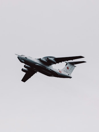 A military aircraft with distinctive radars and aerodynamic design flies through a cloudy sky. The airplane is painted in gray and has recognizable markings on its body, with four powerful jet engines under its wings.