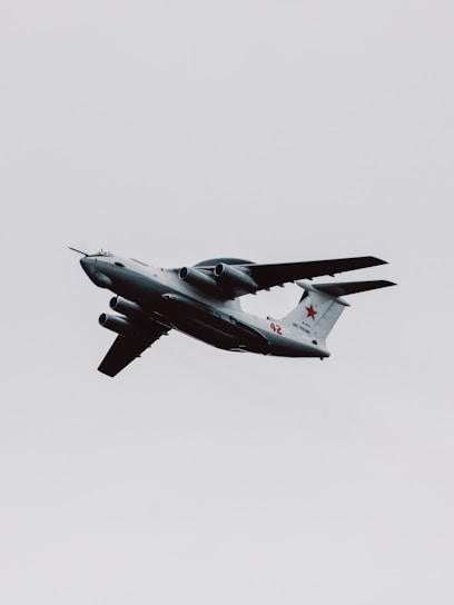A military aircraft with distinctive radars and aerodynamic design flies through a cloudy sky. The airplane is painted in gray and has recognizable markings on its body, with four powerful jet engines under its wings.