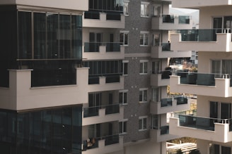 A cluster of modern apartment buildings with large glass balconies and windows, showcasing multiple levels of residential units. The architecture features a combination of light materials and darker window frames, creating a contrast. The balconies appear spacious, and there is greenery visible in the background, suggesting an urban setting with some natural elements.