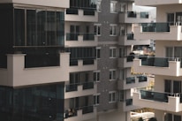 A cluster of modern apartment buildings with large glass balconies and windows, showcasing multiple levels of residential units. The architecture features a combination of light materials and darker window frames, creating a contrast. The balconies appear spacious, and there is greenery visible in the background, suggesting an urban setting with some natural elements.