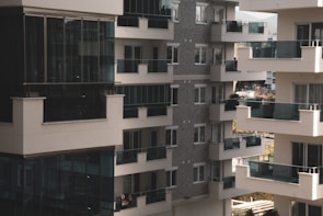 A cluster of modern apartment buildings with large glass balconies and windows, showcasing multiple levels of residential units. The architecture features a combination of light materials and darker window frames, creating a contrast. The balconies appear spacious, and there is greenery visible in the background, suggesting an urban setting with some natural elements.
