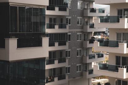 A cluster of modern apartment buildings with large glass balconies and windows, showcasing multiple levels of residential units. The architecture features a combination of light materials and darker window frames, creating a contrast. The balconies appear spacious, and there is greenery visible in the background, suggesting an urban setting with some natural elements.