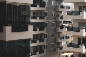 A cluster of modern apartment buildings with large glass balconies and windows, showcasing multiple levels of residential units. The architecture features a combination of light materials and darker window frames, creating a contrast. The balconies appear spacious, and there is greenery visible in the background, suggesting an urban setting with some natural elements.