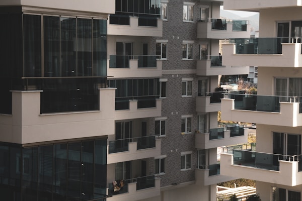 A cluster of modern apartment buildings with large glass balconies and windows, showcasing multiple levels of residential units. The architecture features a combination of light materials and darker window frames, creating a contrast. The balconies appear spacious, and there is greenery visible in the background, suggesting an urban setting with some natural elements.