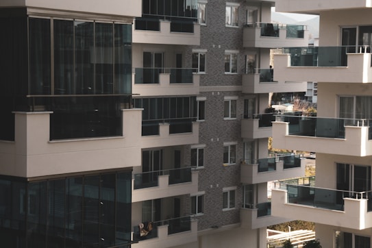 A cluster of modern apartment buildings with large glass balconies and windows, showcasing multiple levels of residential units. The architecture features a combination of light materials and darker window frames, creating a contrast. The balconies appear spacious, and there is greenery visible in the background, suggesting an urban setting with some natural elements.