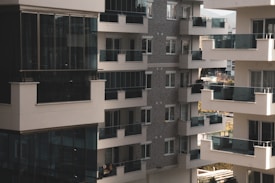 A cluster of modern apartment buildings with large glass balconies and windows, showcasing multiple levels of residential units. The architecture features a combination of light materials and darker window frames, creating a contrast. The balconies appear spacious, and there is greenery visible in the background, suggesting an urban setting with some natural elements.