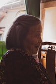 An elderly person with a gentle smile, dressed in a floral-patterned garment, sits in a chair. Warm light filters in, casting soft shadows, and a wooden frame and green fabric are visible in the background.