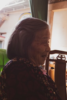 An elderly person with a gentle smile, dressed in a floral-patterned garment, sits in a chair. Warm light filters in, casting soft shadows, and a wooden frame and green fabric are visible in the background.