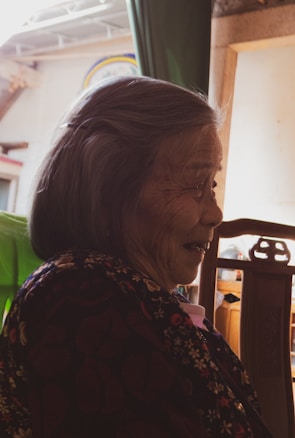 An elderly person with a gentle smile, dressed in a floral-patterned garment, sits in a chair. Warm light filters in, casting soft shadows, and a wooden frame and green fabric are visible in the background.