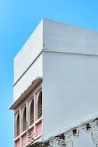 A building facade with a minimalist design featuring a white, cube-like structure. Below the main structure are several arched windows with red lattice detail, partially obscured by climbing greenery. The lower portion of the wall displays weathered brickwork, suggesting age and deterioration.