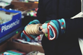 A person wearing colorful knitted gloves holds a roll of twine. The background features a blue box with the word 'Plasencia' and a variety of items that appear to be in a market or store setting.