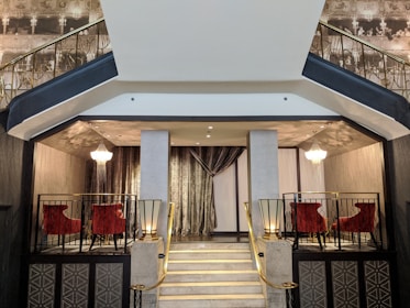 A luxurious interior setting with an elegant staircase leading up to a small seating area. The area is adorned with rich, red velvet chairs and is flanked by ornate chandeliers that provide soft lighting. The walls are decorated with intricate patterns and a silver-toned curtain drapes behind the seating area. The banisters of the staircase are accented with gold details, enhancing the opulent atmosphere.