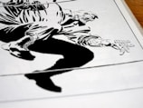 A stylized, high-contrast black and white drawing of a crouching figure with exaggerated features. The illustration appears to be from a comic book, depicting dynamic motion through bold lines and angular shapes. The figure's hand and boot are prominently detailed, capturing a sense of action.