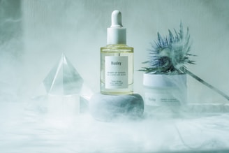 A bottle of Huxley skincare oil is placed on a smooth gray stone, surrounded by a misty atmosphere. To the right, a spiky, bluish thistle flower adds a natural element, and behind the objects is another Huxley skincare product. A transparent geometric crystal structure is placed in the background, contributing to the serene aesthetic.