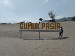 A large, bold sign with the words 'GUMUK PASIR' stands prominently on sandy terrain. A lone person is standing next to the sign, wearing a backpack. In the background, sparse vegetation and a small pavilion structure on a dune can be seen under a bright sky.