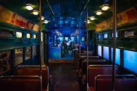 The interior of a vintage bus with rows of brown seats and overhead advertisements. The lighting is dim with a bluish glow, and there are small windows on either side. The back of the bus shows a projection of people, possibly depicting a historical scene.