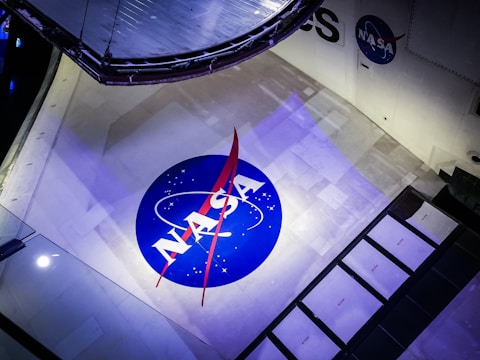The image features a large NASA logo displayed prominently on a spacecraft. The logo is circular with a blue background, white letters, and a red vector symbol. The surface around the logo is a light grey with some panels and mechanical components visible, suggesting the setting is within a space vehicle or exhibit.