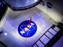 The image features a large NASA logo displayed prominently on a spacecraft. The logo is circular with a blue background, white letters, and a red vector symbol. The surface around the logo is a light grey with some panels and mechanical components visible, suggesting the setting is within a space vehicle or exhibit.