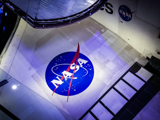 The image features a large NASA logo displayed prominently on a spacecraft. The logo is circular with a blue background, white letters, and a red vector symbol. The surface around the logo is a light grey with some panels and mechanical components visible, suggesting the setting is within a space vehicle or exhibit.
